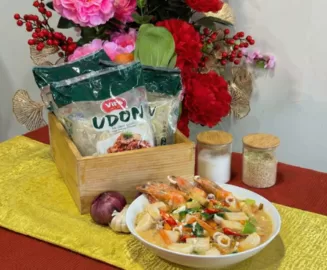 Bowl of Cantonese Fried Udon (Kung Fu style) topped with whole prawns, sliced fishcakes, choy sum and carrots in a glossy garlic-soy gravy, placed beside a wooden box of Vit’s Udon packets, garlic and onion cloves, and two small jars on a gold runner with Chinese New Year backdrop