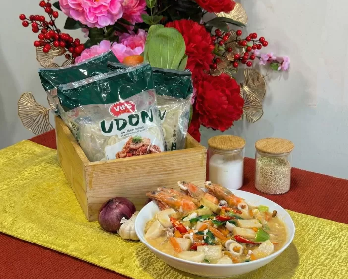 Bowl of Cantonese Fried Udon (Kung Fu style) topped with whole prawns, sliced fishcakes, choy sum and carrots in a glossy garlic-soy gravy, placed beside a wooden box of Vit’s Udon packets, garlic and onion cloves, and two small jars on a gold runner with Chinese New Year backdrop